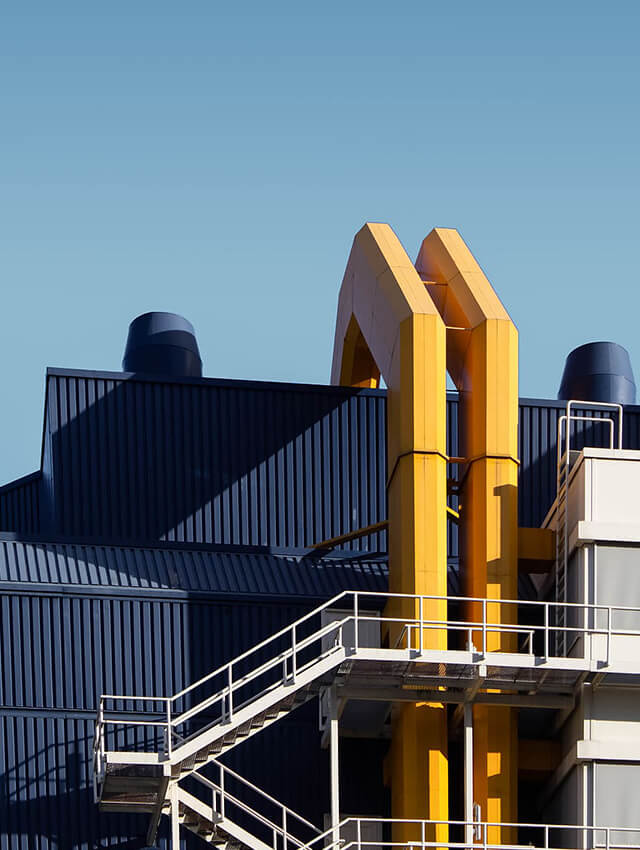 services-industrial-painting Industrial building exterior with yellow structural pipes and blue corrugated metal walls under clear sky